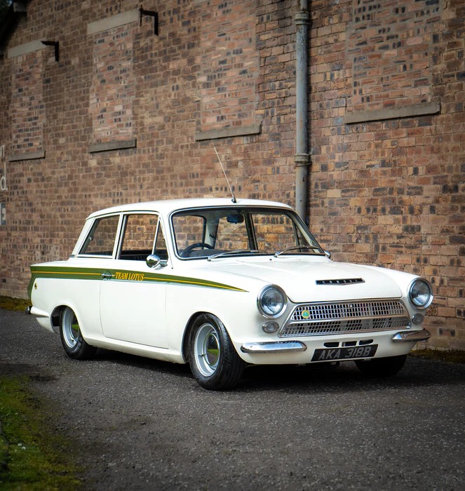 White 1964 Ford Lotus Cortina MK1 with green stripes parked in front of brick building