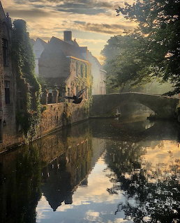 Canal in Bruges with historic buildings and trees reflected in still water under evening sky
