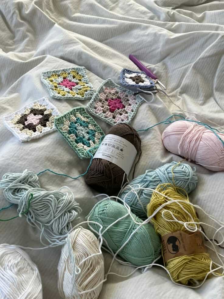 Assorted pastel yarn balls and crocheted granny squares on a white cloth surface