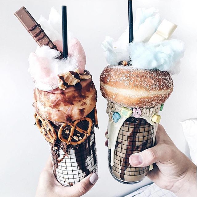Two hands holding decorated milkshakes topped with donuts, cotton candy, pretzels, and chocolate
