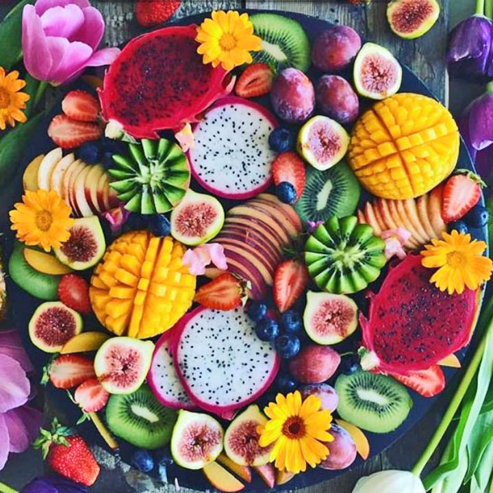 Overhead view of a colorful fruit tray with mango, kiwi, figs, strawberries, bananas, dragon fruit, and flowers