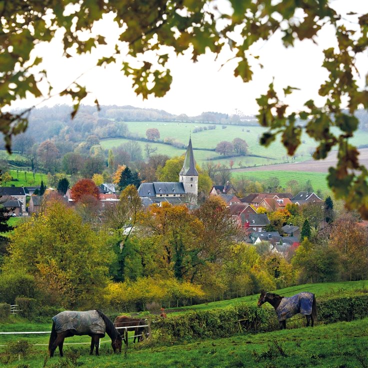 Village with church surrounded by green countryside hills, framed by tree branches with grazing horses in foreground