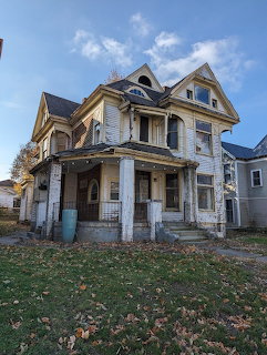 Old Victorian house with peeling paint, front porch, and grassy yard beneath a blue sky.