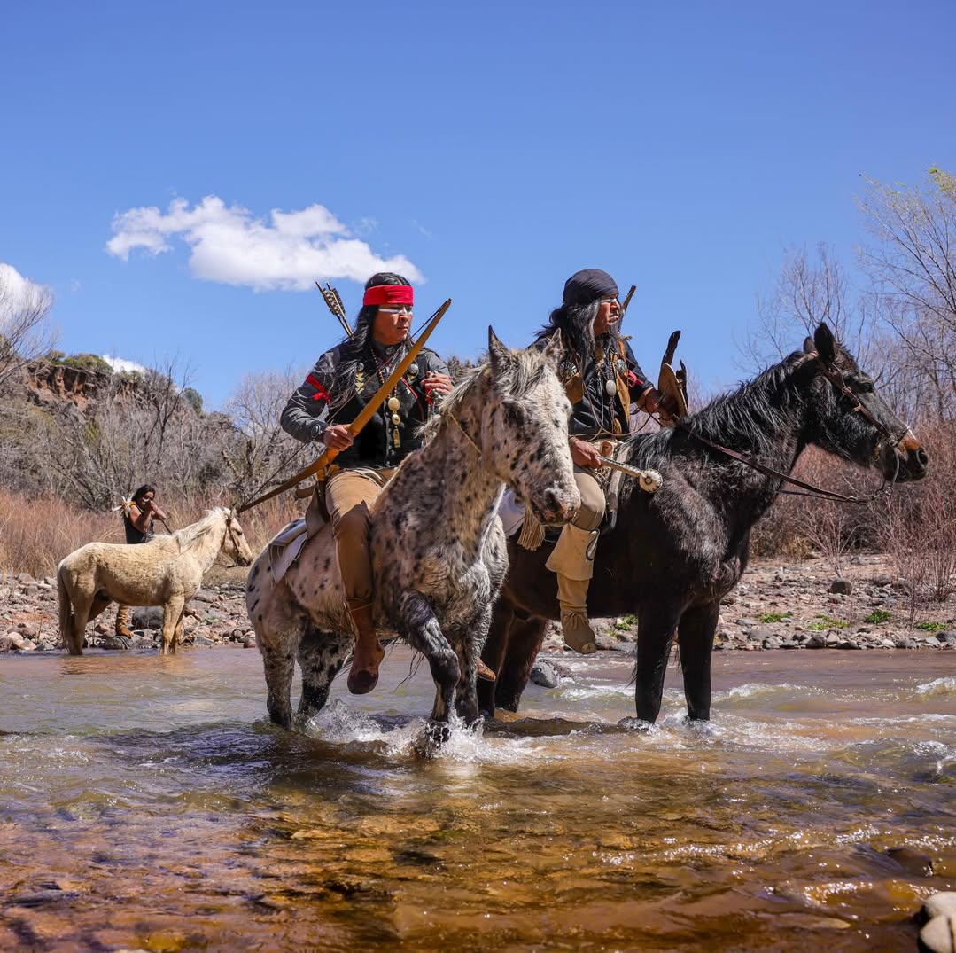 Two riders on horses crossing a shallow river with a dog nearby under a blue sky