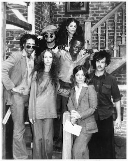Black and white photo of seven people posing together on brick steps in 1970s clothing