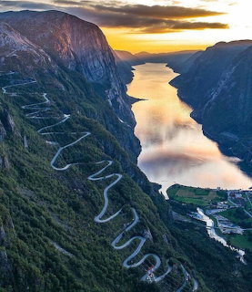 Winding switchback road descending a steep green mountainside to a fjord at sunset