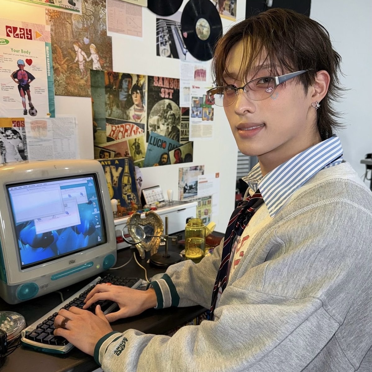 Person with glasses using a computer at a desk in a poster-covered room