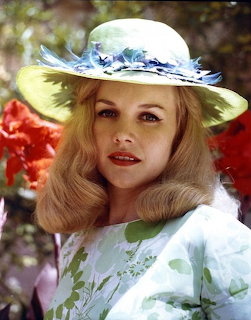 Blonde woman in white hat with blue feathers and floral dress posed among red flowers