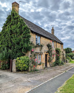 Ivy-covered Cotswold stone cottage with climbing roses on a village street under cloudy sky