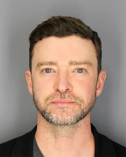 Mugshot of a man with short brown hair, blue eyes, and a beard wearing a black shirt