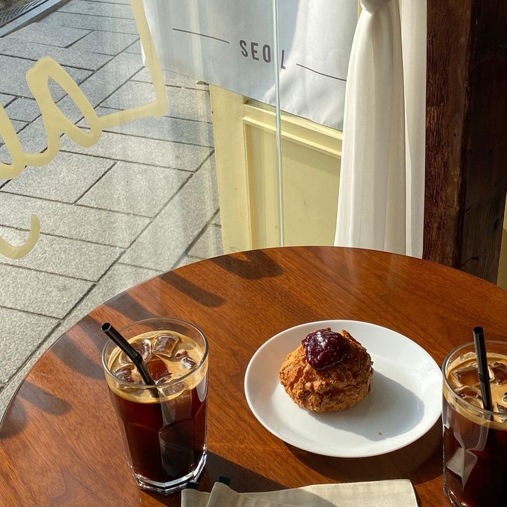 Two glasses of iced coffee and a pastry on a round wooden cafe table with sunlight and tiled floor visible
