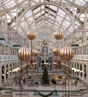 Large Christmas ornaments hanging in a glass-roofed indoor atrium above a decorated tree