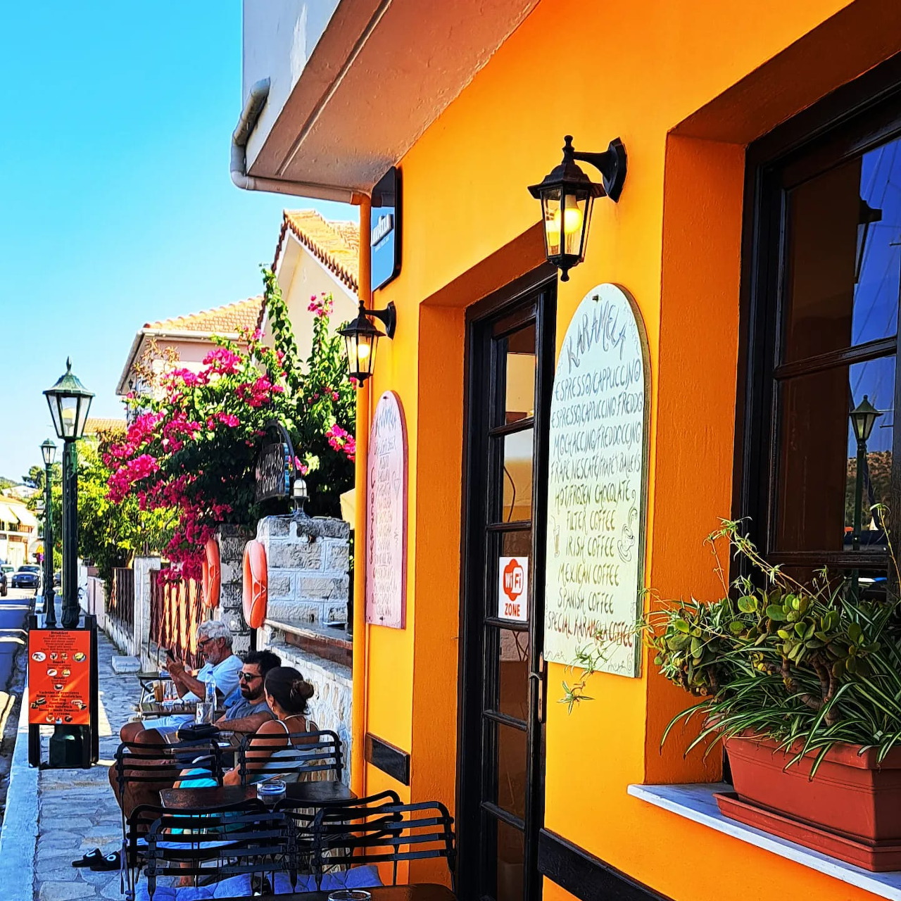 Outdoor cafe tables beside a bright orange building on a sunlit narrow street with flowers and lanterns