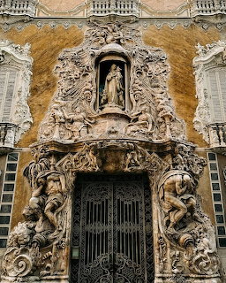 Ornate baroque church entrance with carved stone figures and religious statue in decorative niche above iron gate