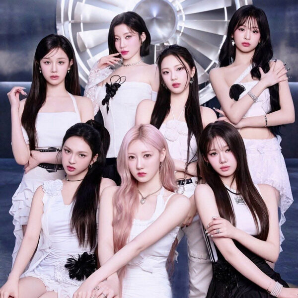 Seven young women in white outfits posing together against a metallic studio backdrop