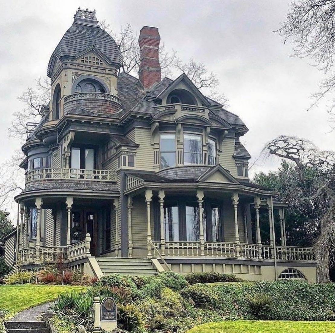 Large Victorian mansion with turret, wrap porch, ornate details, green lawn and bare trees