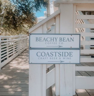 White directional sign for Beachy Bean Coffee Co. and Coastside establishments with white picket fence and tree