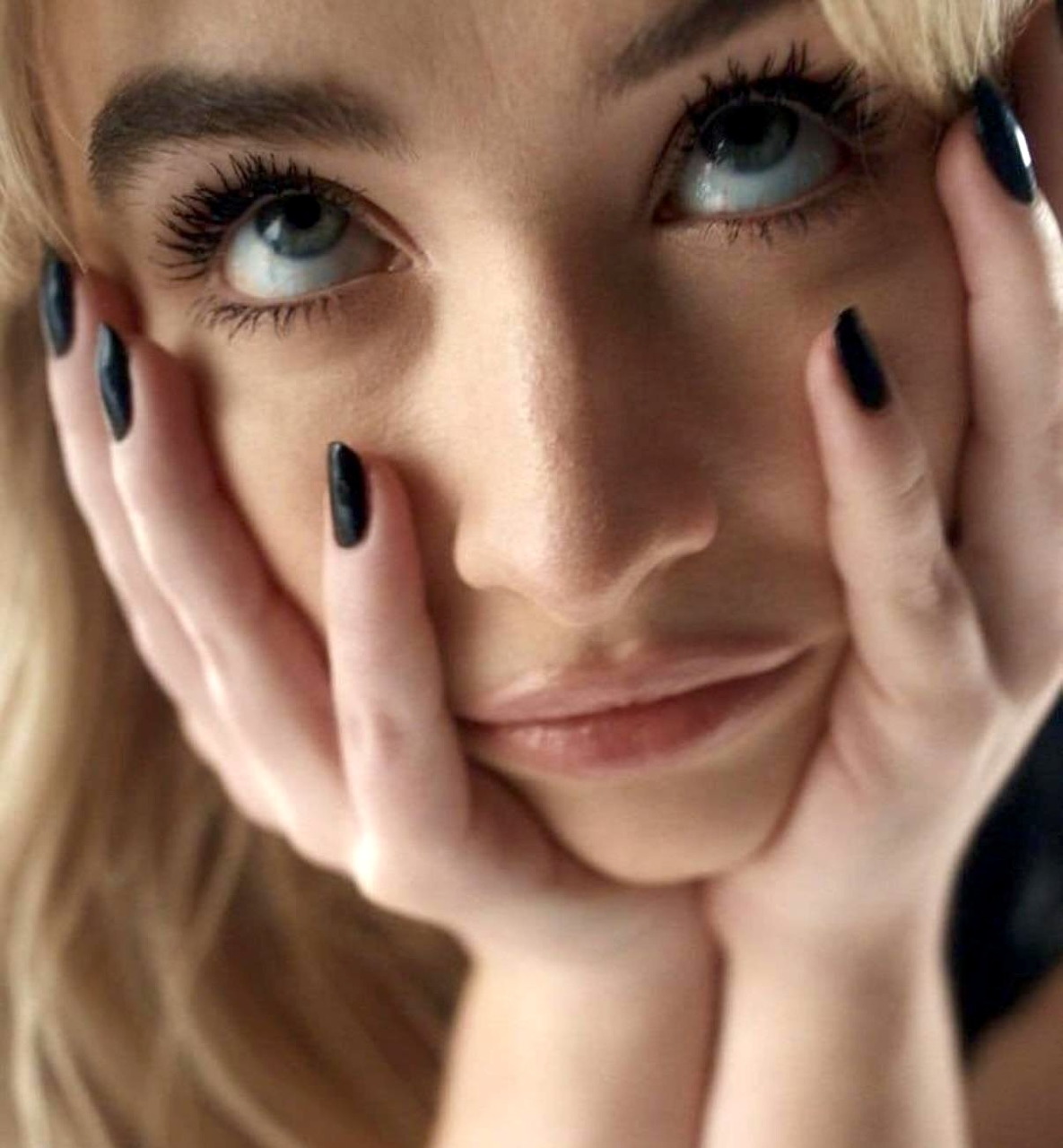 Close-up of a blonde woman with blue eyes and black nail polish, hands cupping her face