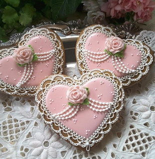 Three pink heart-shaped decorated cookies with lace icing and rose decorations on a lace cloth