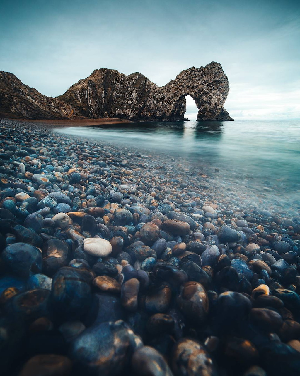 Natural stone arch formation on a pebble beach with calm sea and cliffs under soft blue sky