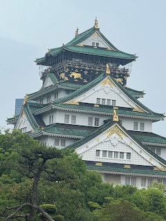 Osaka Castle in Japan with green rofs, gold decorations, and surrounding trees under cloudy sky