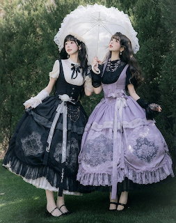 Two women in gothic lolita dresses, one black and one lavender, posing with a white parasol outdoors