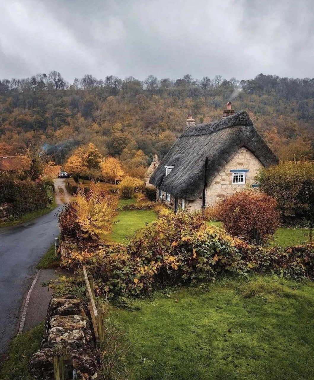 Stone thatched cottage with green lawn surrounded by autumn trees and a winding country road