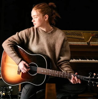 Sadie Sink sitting indoors and playing an acoustic guitar near a piano