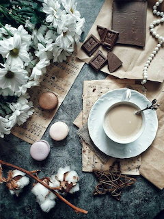 Flatlay with coffee cup, chocolate bars, white flowers, macarons, sheet music, and pearls on grey surface