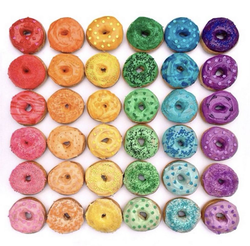 Grid of glazed donuts in rainbow colors with sprinkles arranged in six columns from red through orange, yellow, green, blue to purple