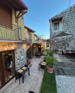 Stone-paved village courtyard between rustic buildings with outdoor tables, chairs, and potted plants