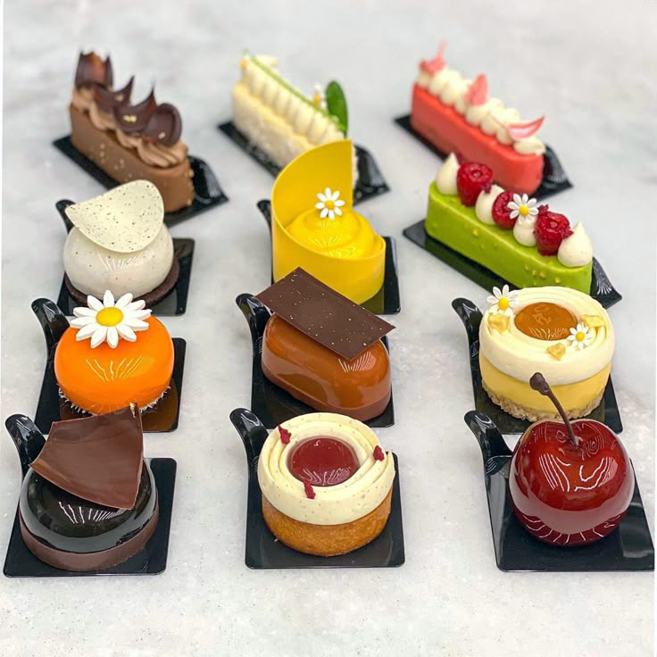 Twelve miniature gourmet desserts arranged in rows on black slate plates with colorful glazes and garnishes