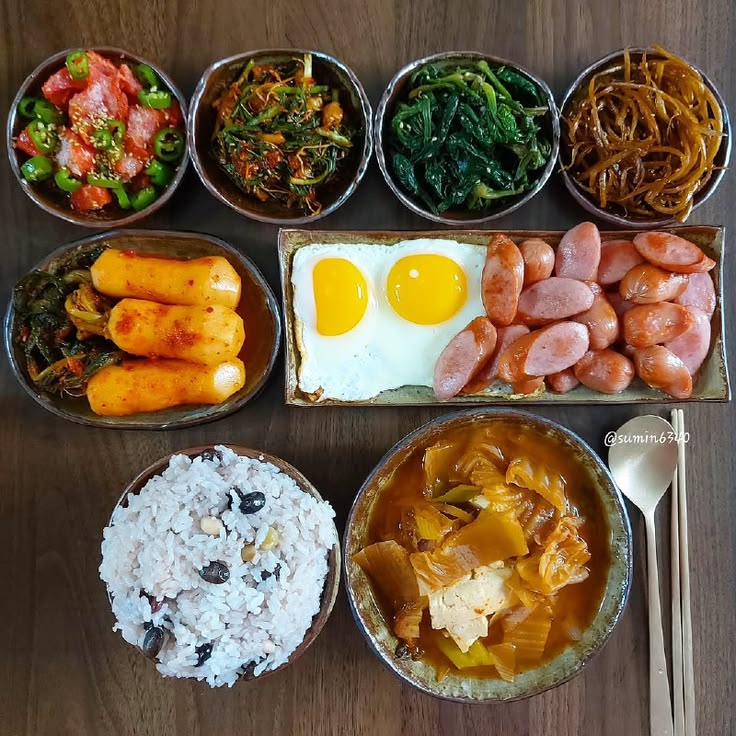 Overhead view of Korean meal with side dishes, eggs, sausages, rice, and stew in various bowls on wooden table