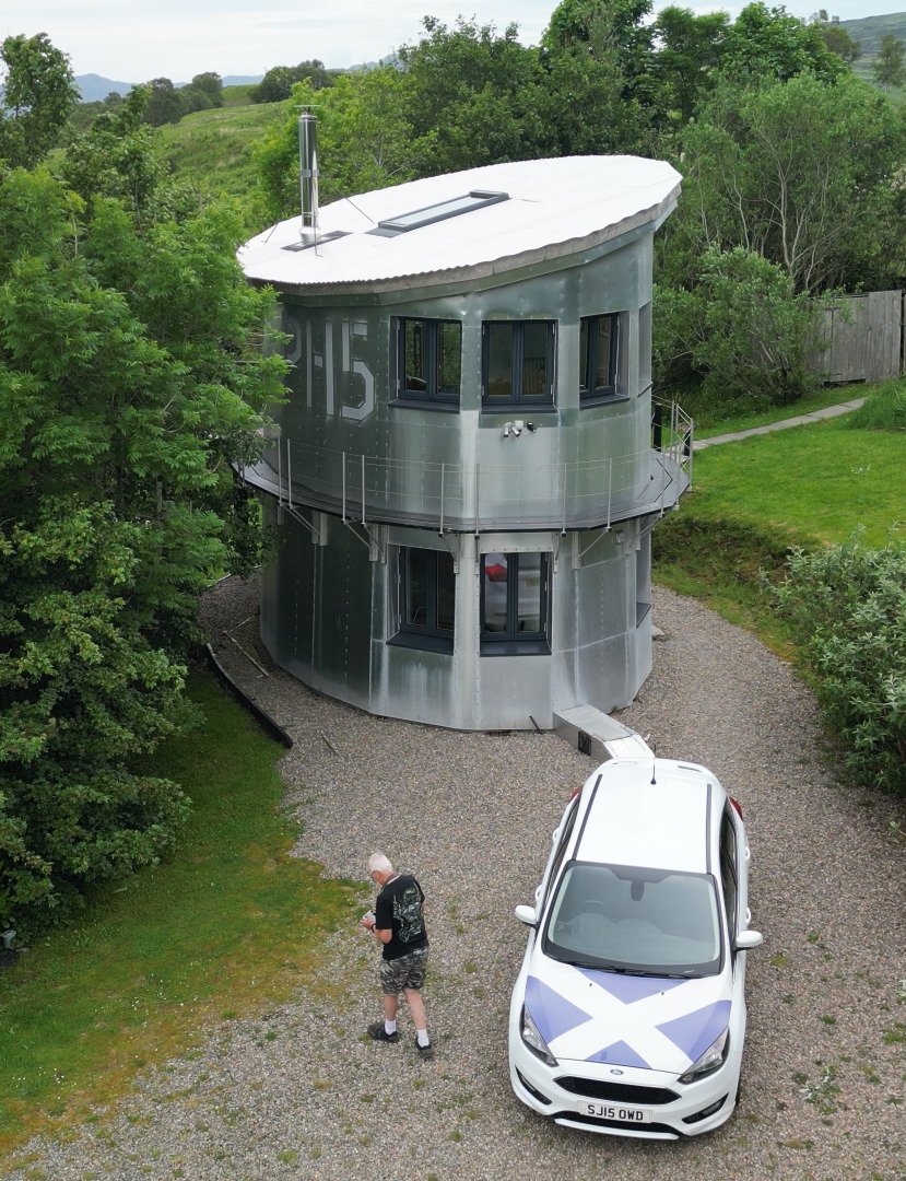 Aerial view of cylindrical gray house with white curved roof, person and white car on gravel drive, surrounded by green trees