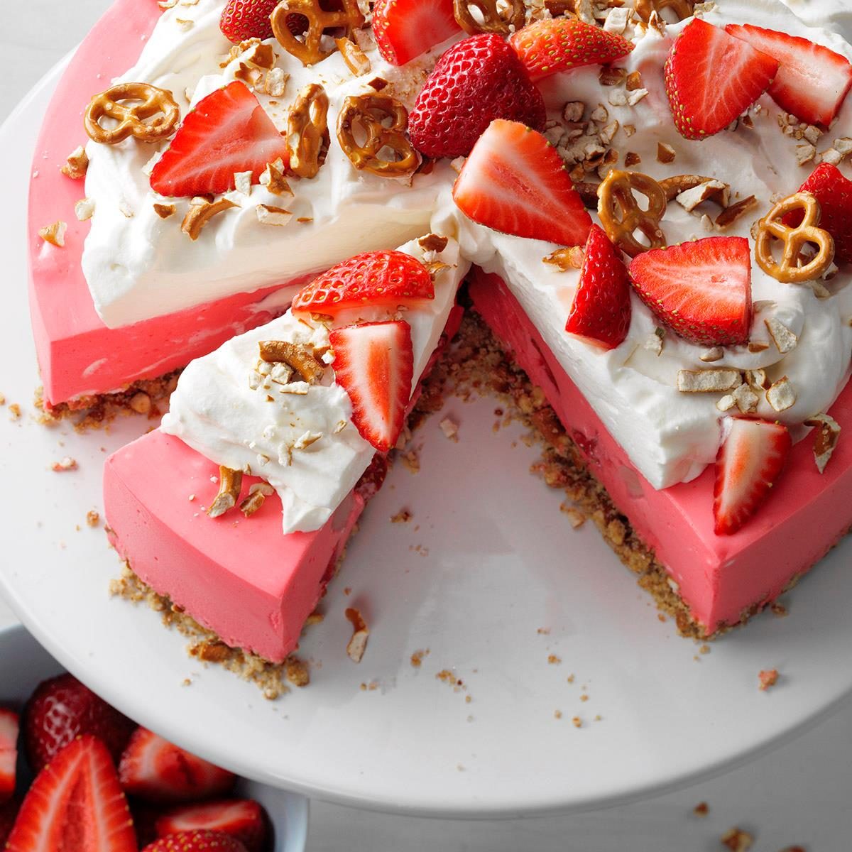 Pink strawberry pretzel dessert cake with whipped cream topping, fresh strawberries, and crushed pretzels on white plate