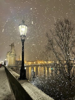 Glowing street lamp on snowy Prague bridge at night with castle towers and bare trees in background