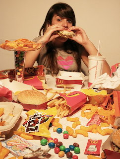 Woman biting pizza slice at table covered with fast food, burgers, fries, donuts, candy and soda