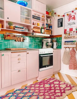 Colorful kitchen with pink cabinets, turquoise tiles, open shelves, and patterned rug on checkered floor