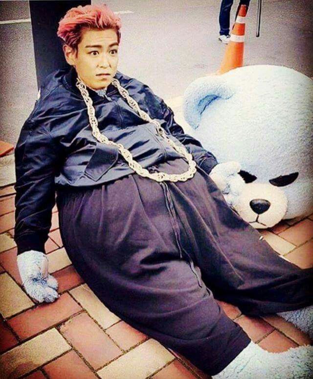 Young man with pink hair and chain necklace sitting on brick floor next to a giant white stuffed polar bear