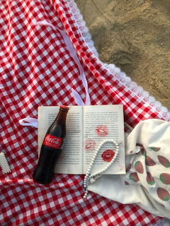Red gingham blanket on sand with open book, cola bottle, pearl necklace, and red lipstick kiss marks