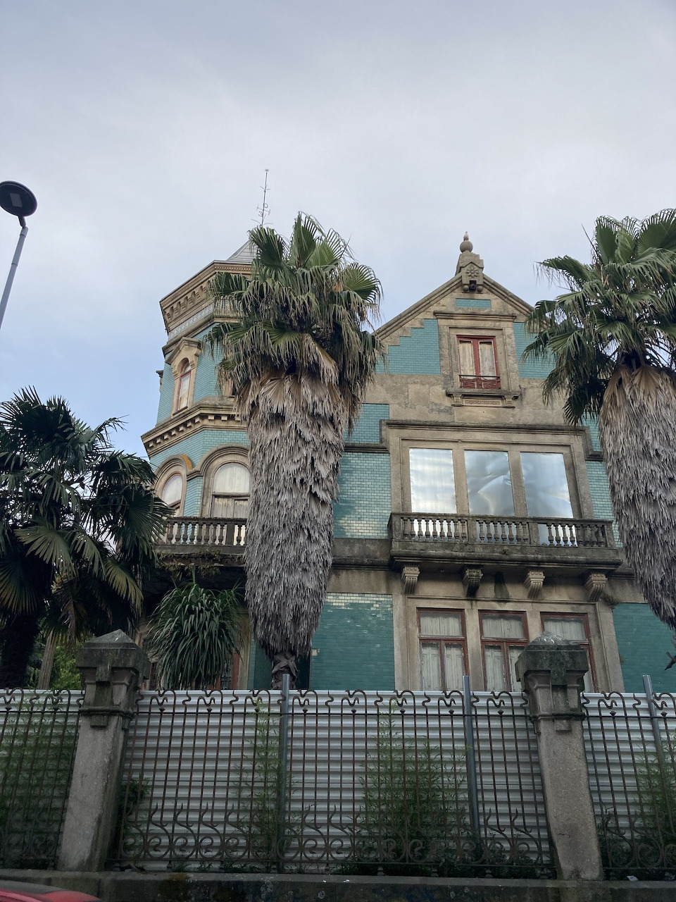 Weathered green Victorian mansion with palm trees, balconies, and a decorative tower against a grey sky