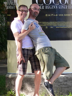 Two men in t-shirts and shorts posing together outdoors in front of a wall