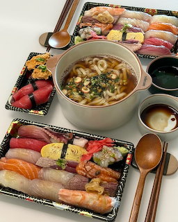 Tabletop spread with sushi trays, noodle soup pot, bowls, and wooden spoons