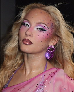 Blonde woman with pink glitter face makeup and purple earring looking at camera