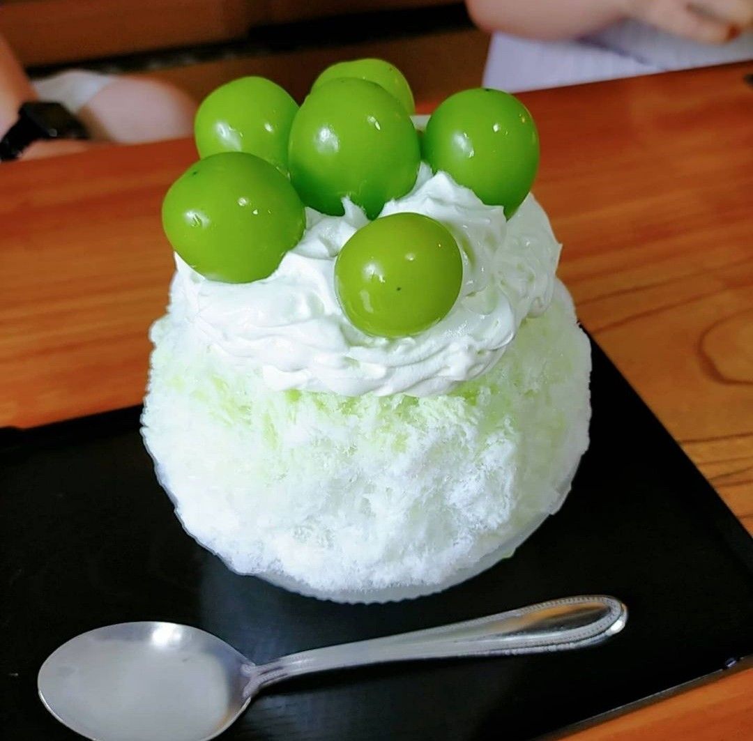 Shaved ice mound with white cream and green grape balls on top, served on black square plate with spoon