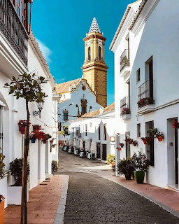 Narrow cobblestone street with white whitewashed buildings and a church tower under blue sky