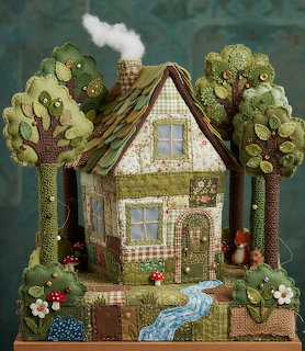 Handcrafted felt cottage with green patchwork roof surrounded by felt trees, mushrooms, and a small stream