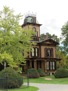 Victorian-style house with a tower, surrounded by trees, shrubs, and a green lawn
