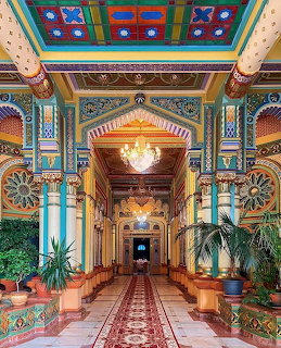 Elaborate hotel hallway with decorated ceiling, ornate columns, chandelier, and red patterned carpet