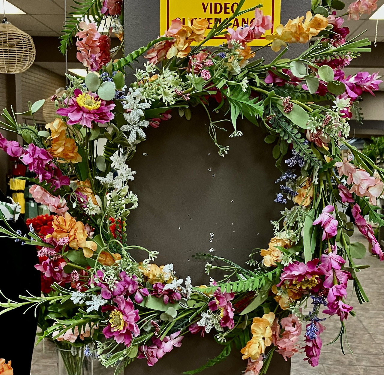 Circular wreath with colorful flowers including pink, white, orange blooms on dark background in garden center
